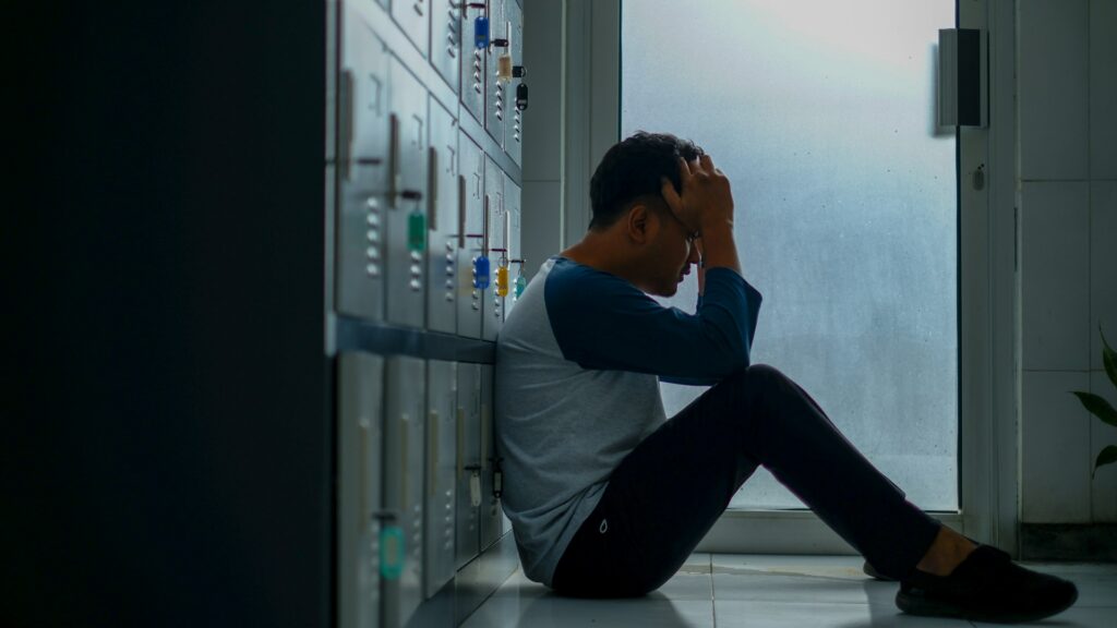 A man sitting on the floor of a locker room, looking worried